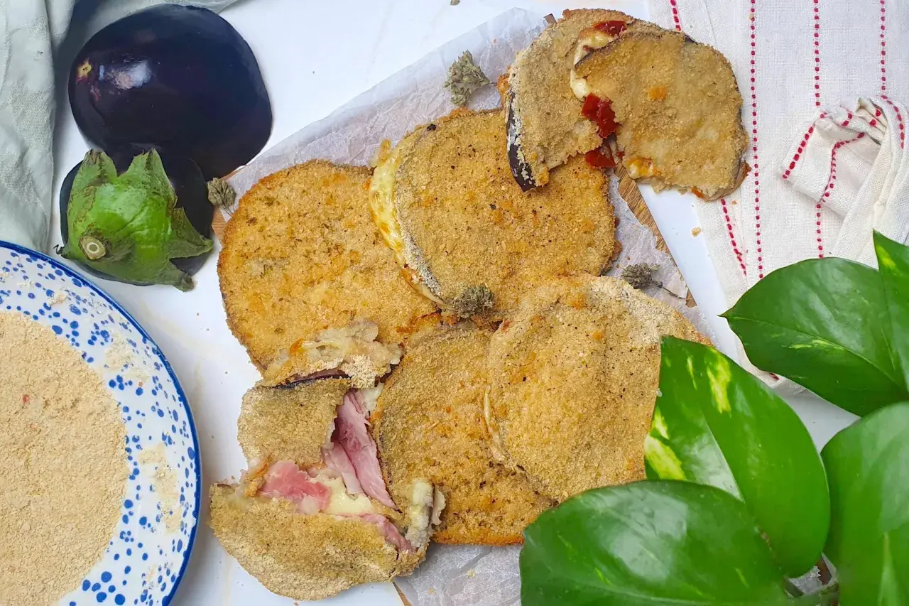 Cordon bleu di melanzane impanati e fritti, ripieni di prosciutto e formaggio filante. Accanto, melanzane fresche e pangrattato.