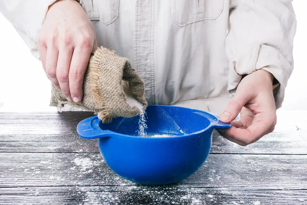 Mani versano zucchero da un sacchetto di juta in una ciotola blu, preparando ingredienti per pasta di zucchero.