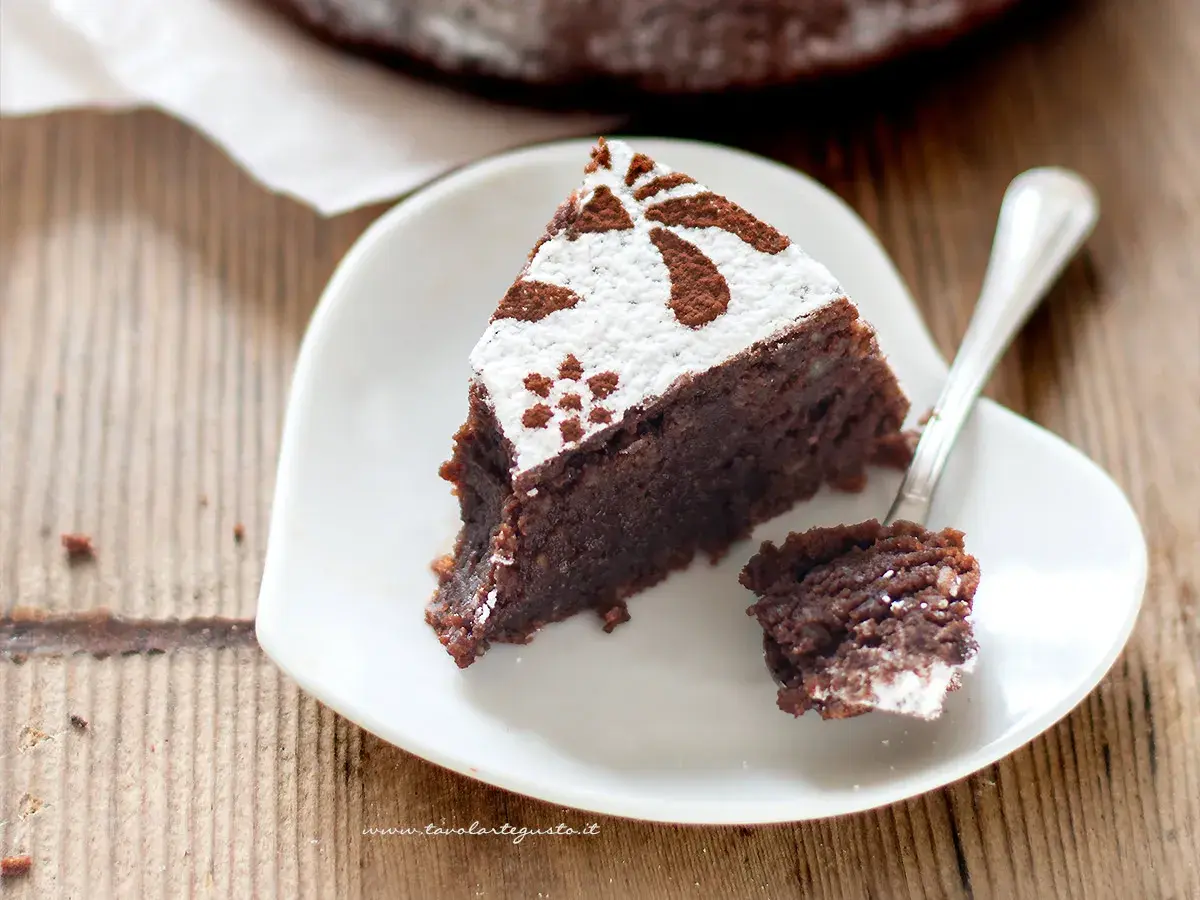 Una fetta di torta caprese bimby, soffice e al cioccolato, decorata con zucchero a velo e cacao, su un piatto bianco a forma di cuore.