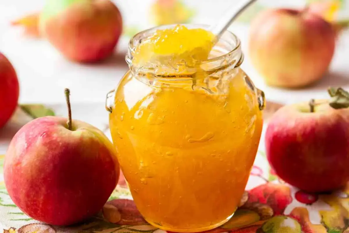 Un barattolo di marmellata di mele dorata, con una cucchiaiata che ne assaggia la dolcezza, circondato da mele rosse fresche.