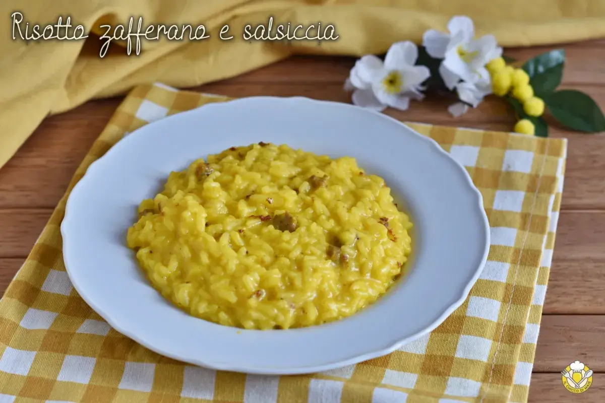 Un cremoso risotto salsiccia e zafferano, servito su un piatto bianco, con fiori gialli e bianchi sullo sfondo.