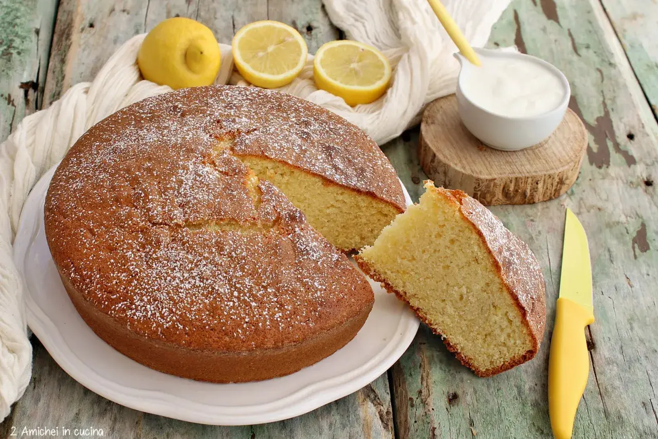 Deliziosa torta per colazione, soffice e profumata al limone, spolverata di zucchero a velo, con una fetta tagliata e una ciotolina di yogurt.