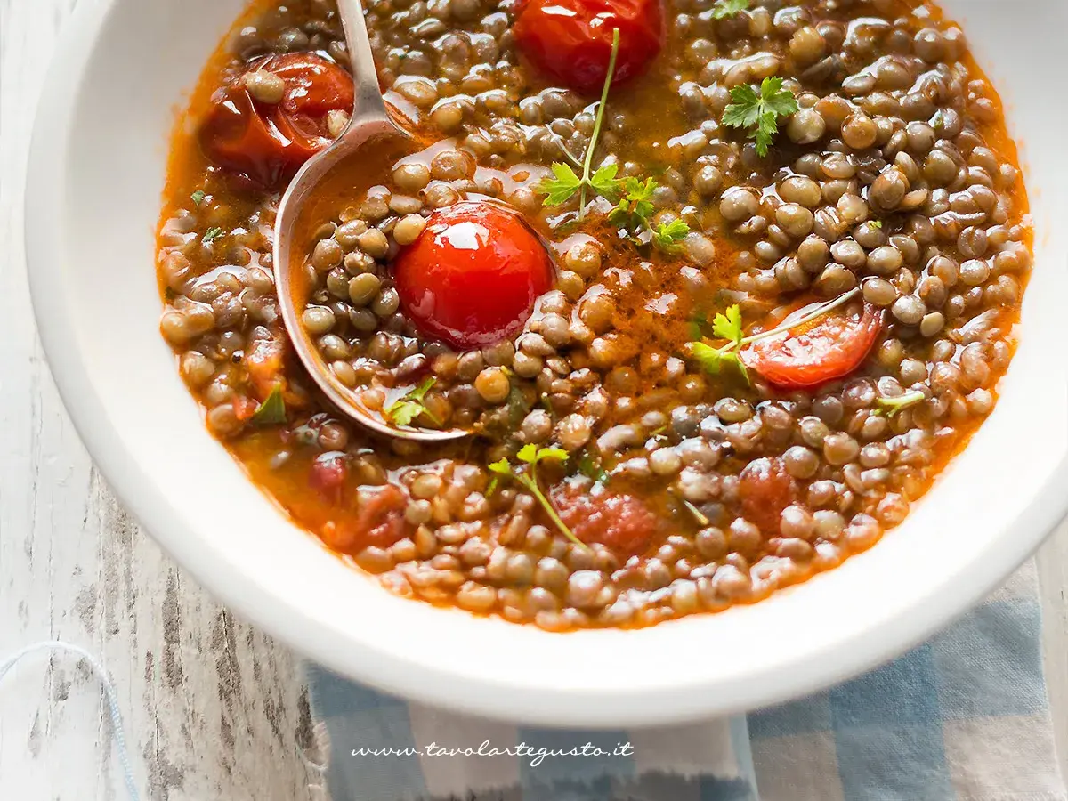 Una ciotola di zuppa di lenticchie fumante, con pomodorini e prezzemolo fresco. Un cucchiaio raccoglie una porzione generosa di questo piatto confortante.