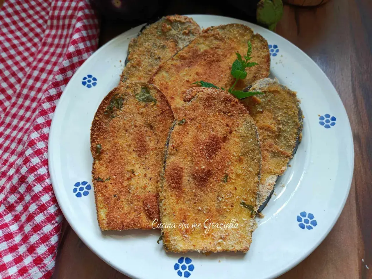 Fette dorate di cotoletta di melanzane impanate, guarnite con menta fresca, su un piatto bianco con decori blu.