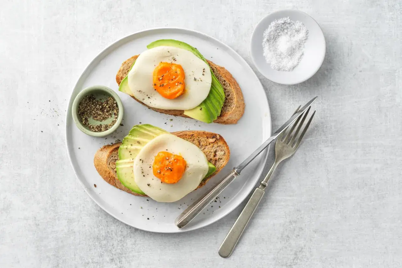 Due fette di pane tostato con avocado, formaggio e un occhio di bue, condite con pepe.