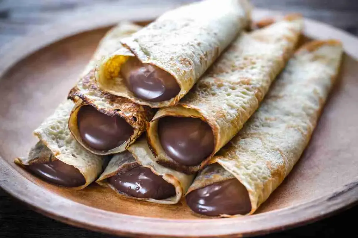 Un mucchio di crepes arrotolate, ripiene di golosa Nutella, su un piatto di legno.