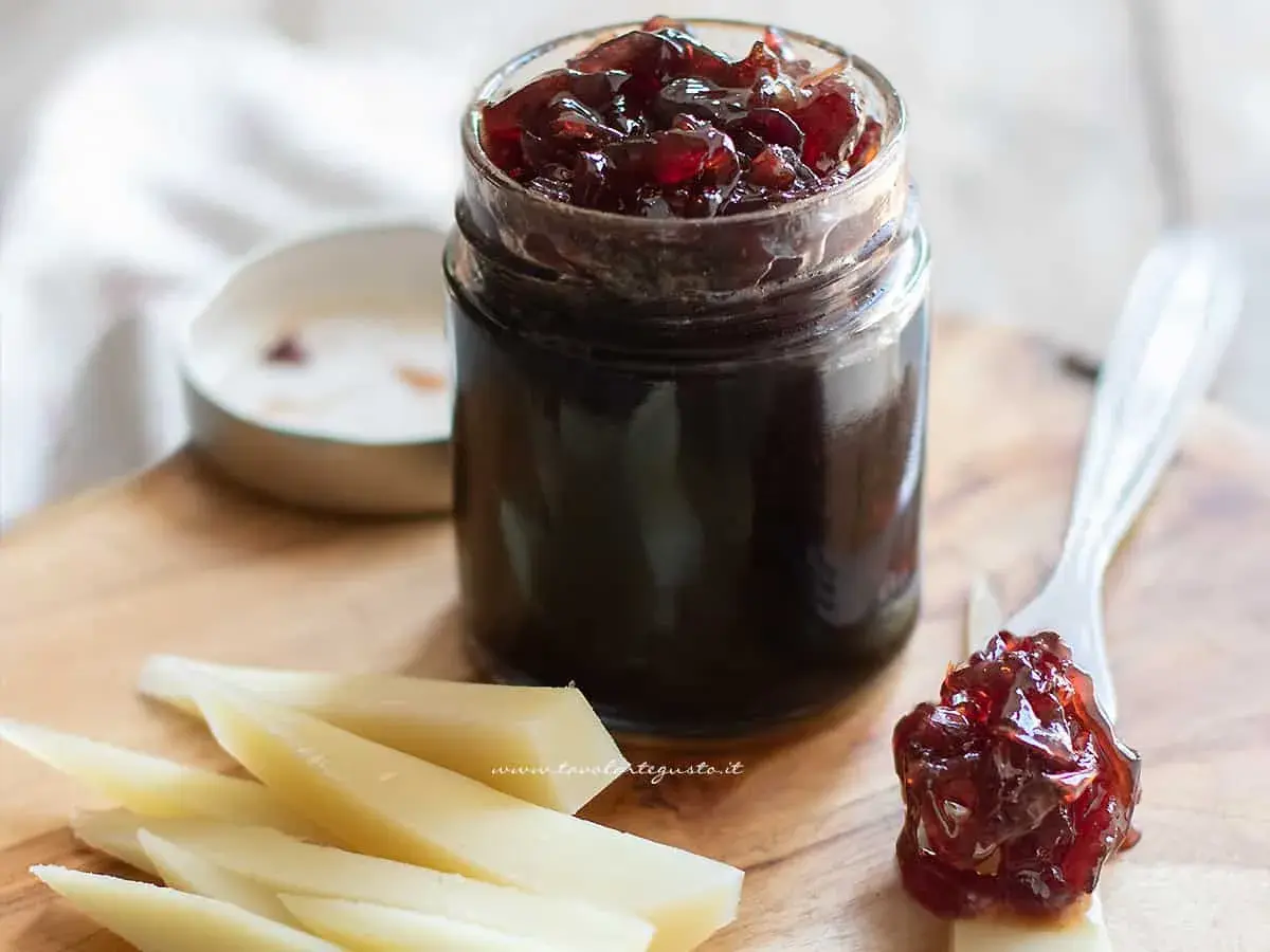 Un vasetto di marmellata di cipolle rosse, con un cucchiaino che ne raccoglie una porzione, accanto a fette di formaggio su un tagliere di legno.