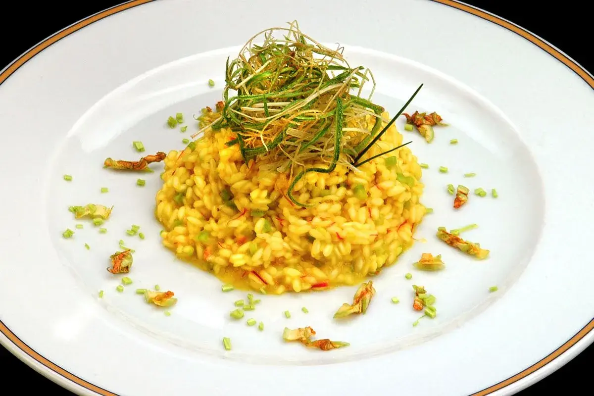 Risotto giallo intenso con fiori di zucca e zafferano, guarnito con erbe croccanti.