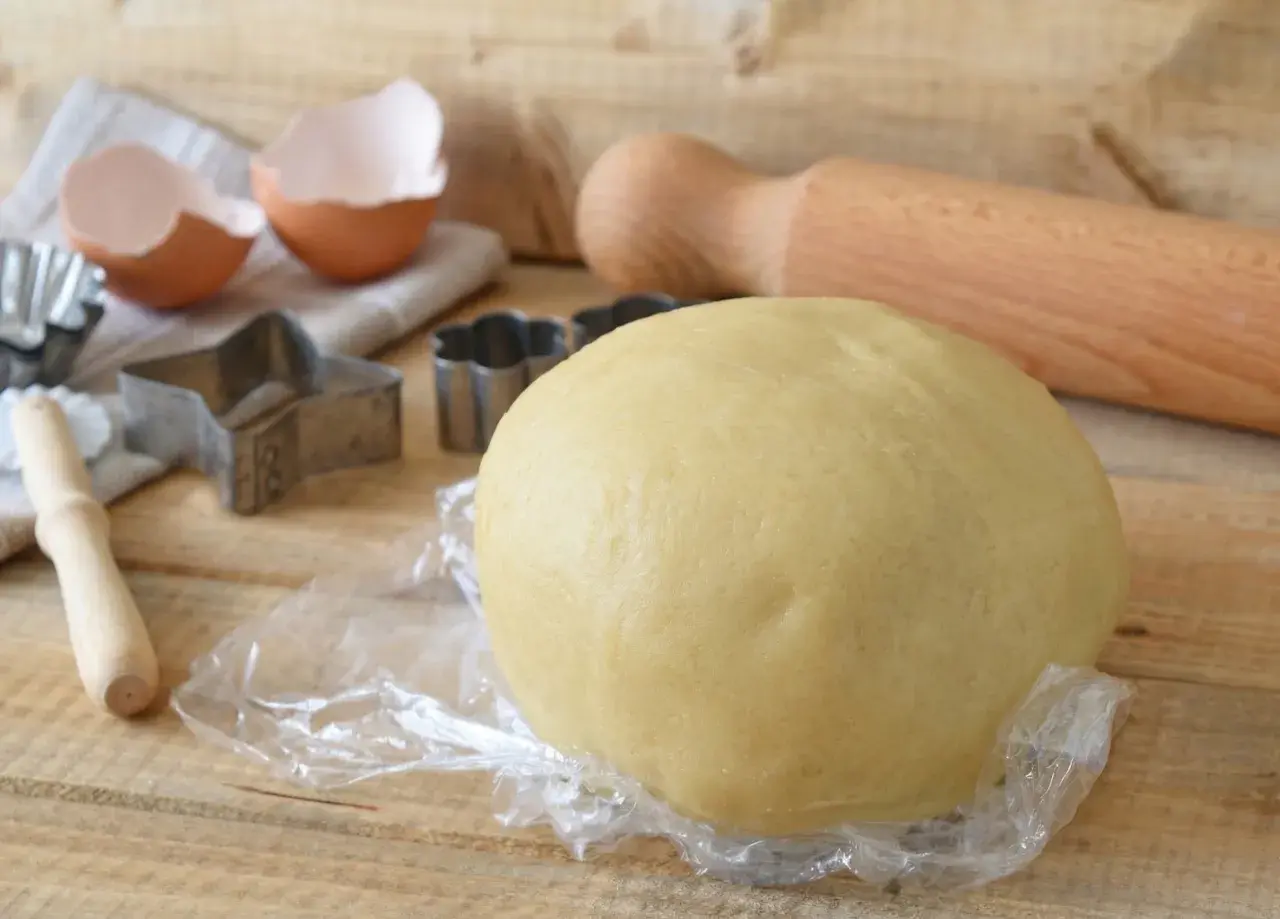 Impasto per crostata pronto, con stampini, uovo rotto e mattarello. La pasta frolla friabile per crostata è perfetta per dolci fatti in casa.