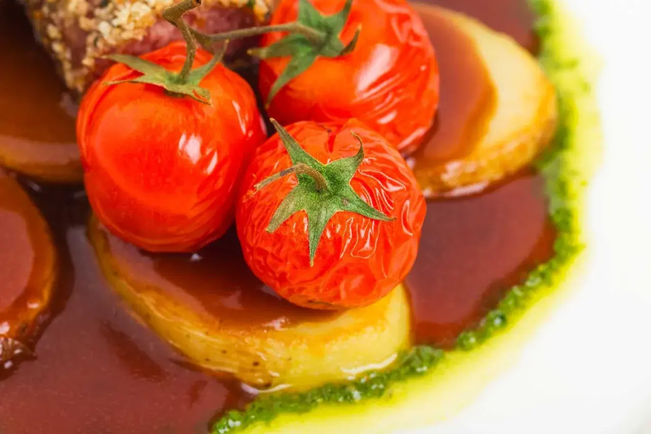 Pomodori alla griglia succosi su patate, immersi in una salsa scura e guarniti con un filo di pesto verde.