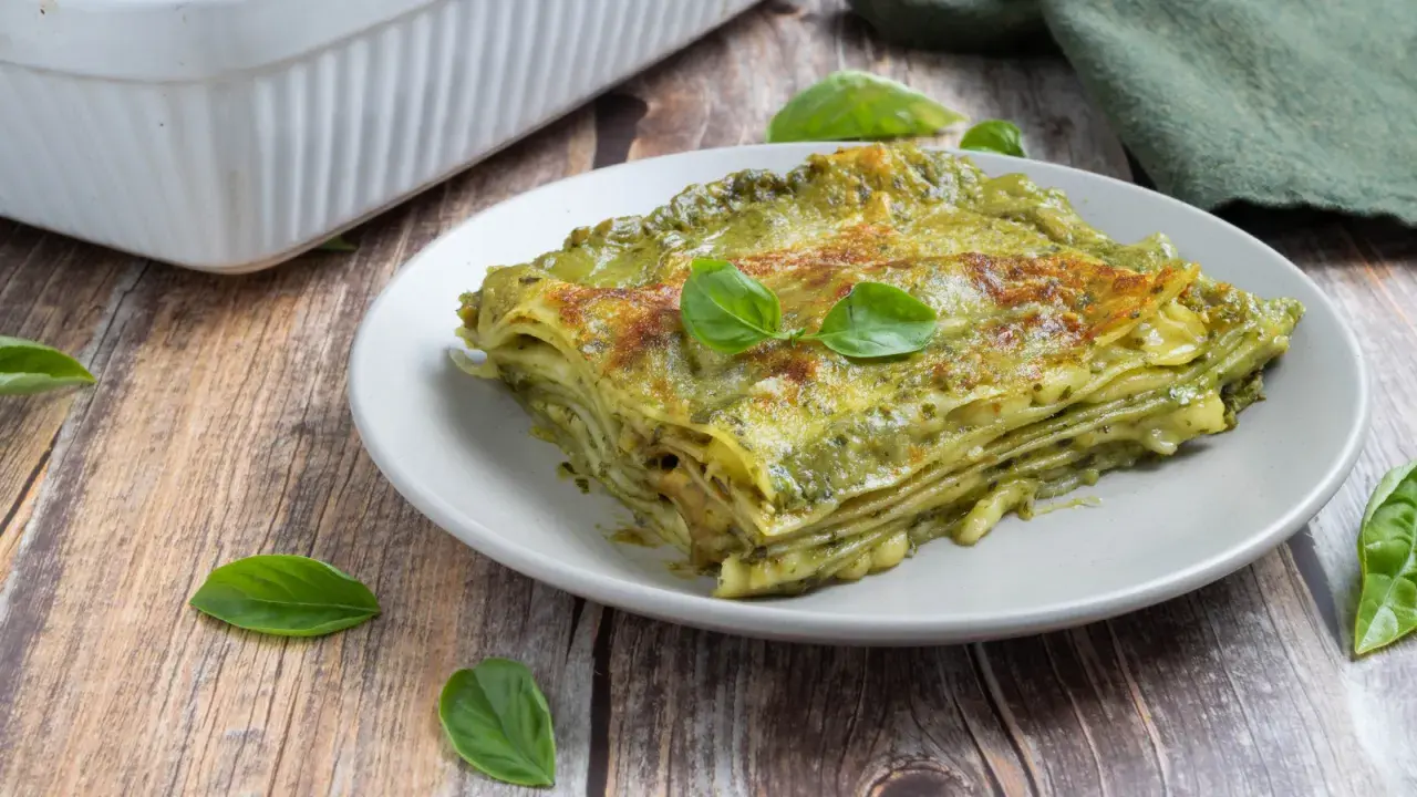 Una fetta di lasagne al pesto, stratificata e dorata, guarnita con basilico fresco su un piatto grigio.