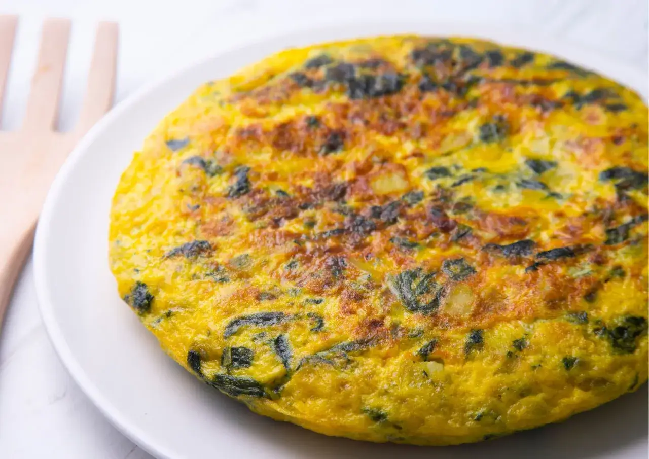Frittata di spinaci dorata e soffice su un piatto bianco, con un utensile in legno sullo sfondo.