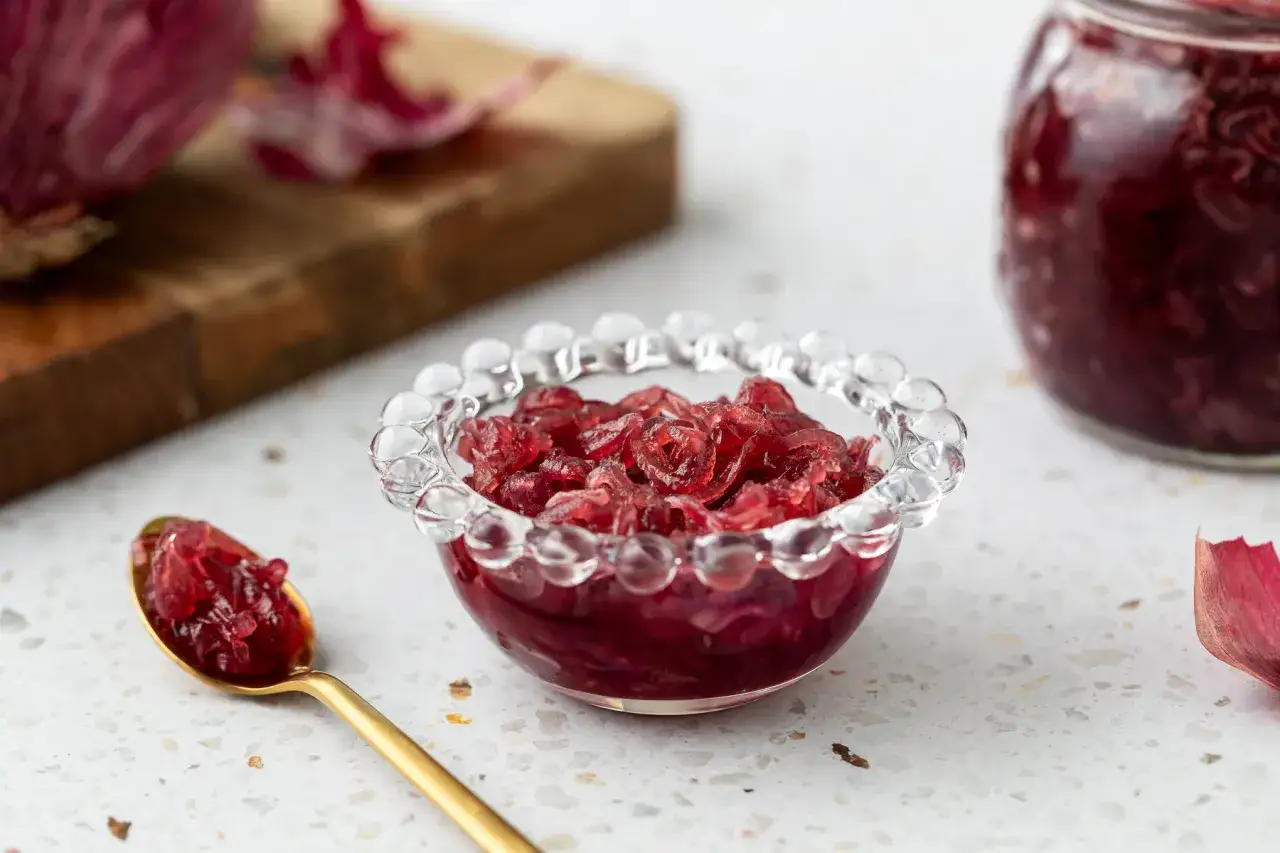 Una ciotola di vetro trasparente piena di marmellata di cipolle rosse, con un cucchiaino dorato e un barattolo sullo sfondo.