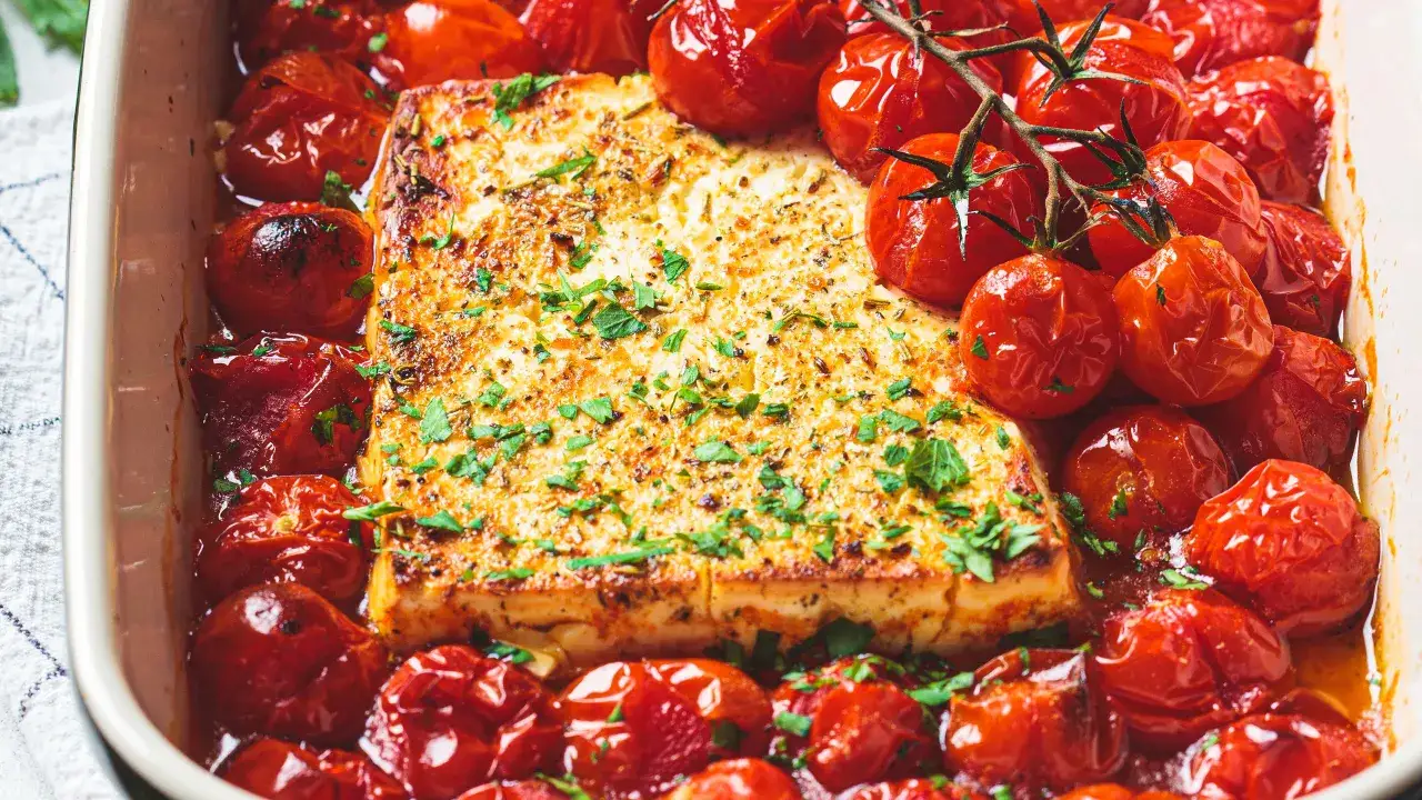 Un blocco di feta al forno, dorato e cosparso di erbe, circondato da pomodorini ciliegino arrostiti in una teglia.