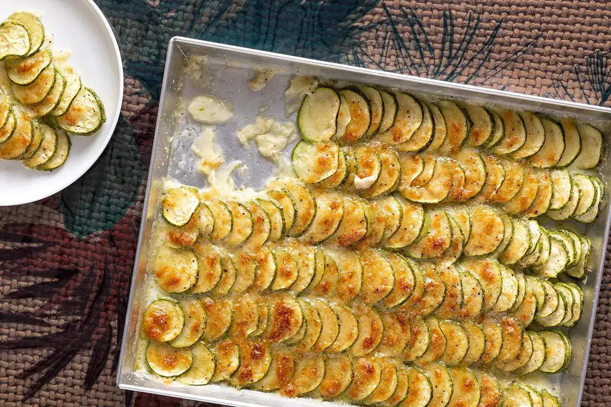 Fette di zucchine al forno, dorate e filanti, disposte in un vassoio e su un piatto bianco.