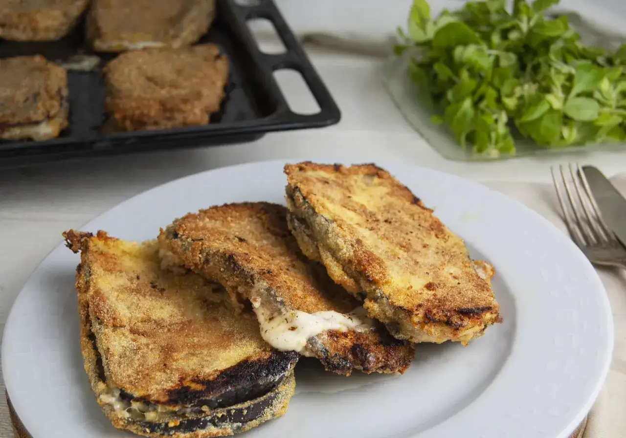 Tre cordon bleu di melanzane dorati e filanti su un piatto bianco, con una teglia e insalata fresca sullo sfondo.
