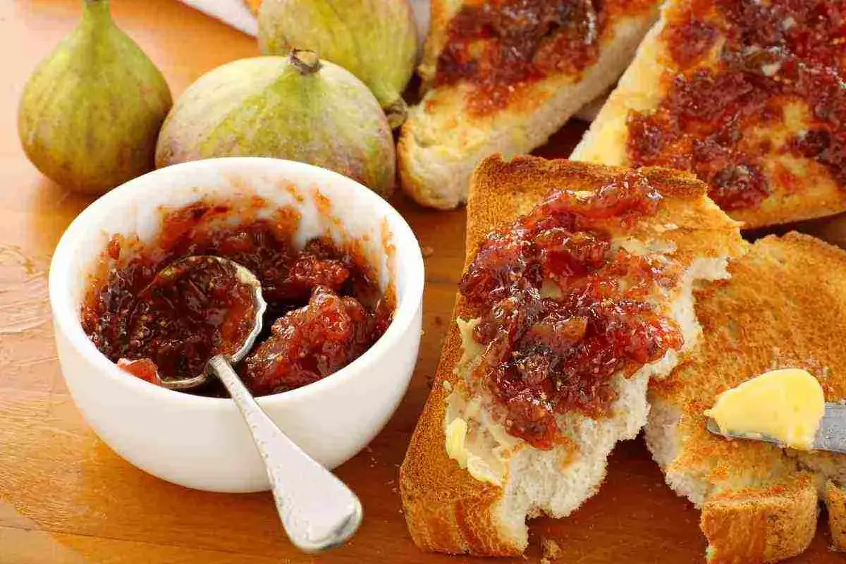 Fette biscottate con composta di fichi, un cucchiaino in una ciotola e fichi freschi sullo sfondo.