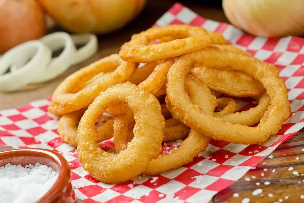 Anelli di cipolla dorati e croccanti, preparati con una pastella per fritti perfetta, serviti su carta a quadri rossi con sale grosso.