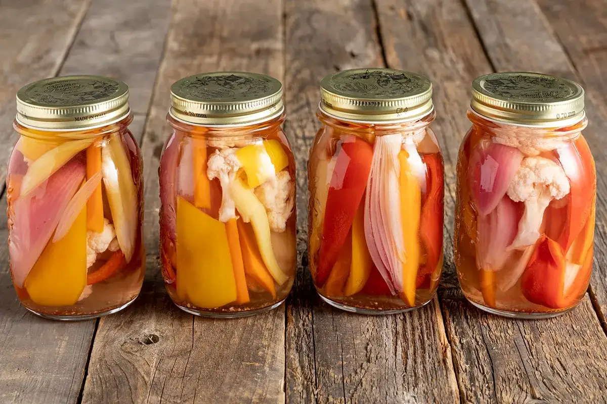 Quattro barattoli di giardiniera fatta in casa, con verdure colorate come peperoni, carote e cavolfiore, conservate in aceto.