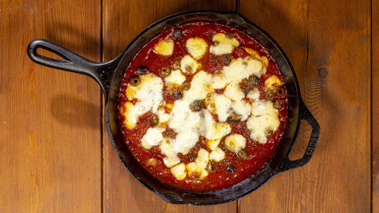 Pollo alla pizzaiola in padella, con pomodoro, olive e mozzarella filante, su un tavolo di legno.