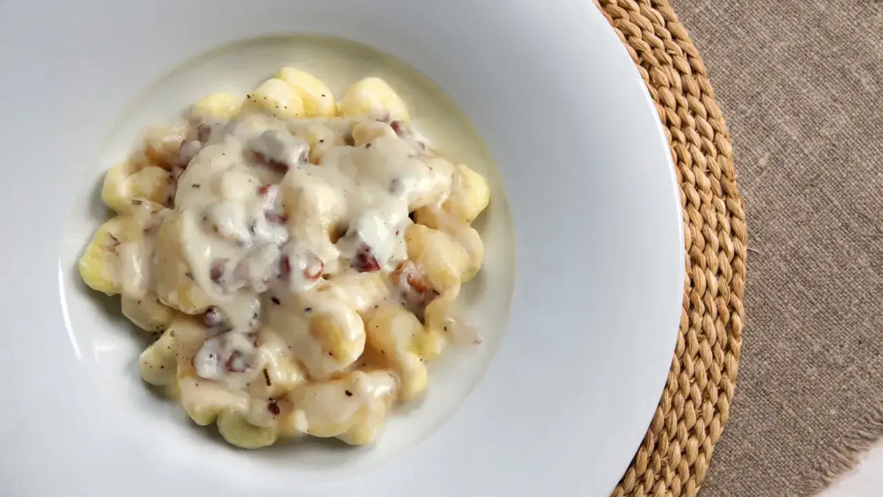 Gnocchi al gorgonzola cremosi, con pezzetti di pancetta croccante, serviti in un piatto bianco su un runner di juta.