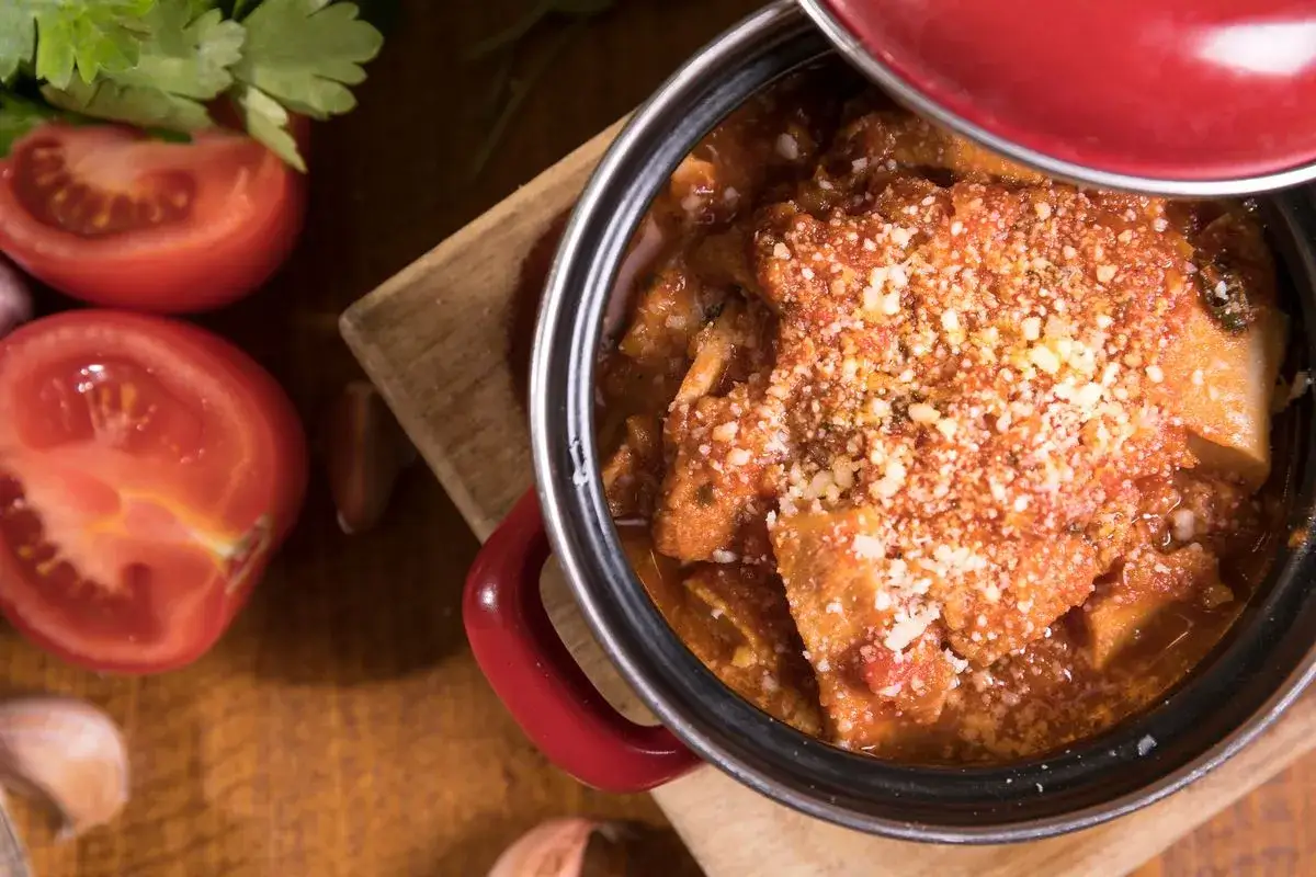 Trippa alla parmigiana fumante in un pentolino rosso, con pomodori freschi e aglio sul tagliere.