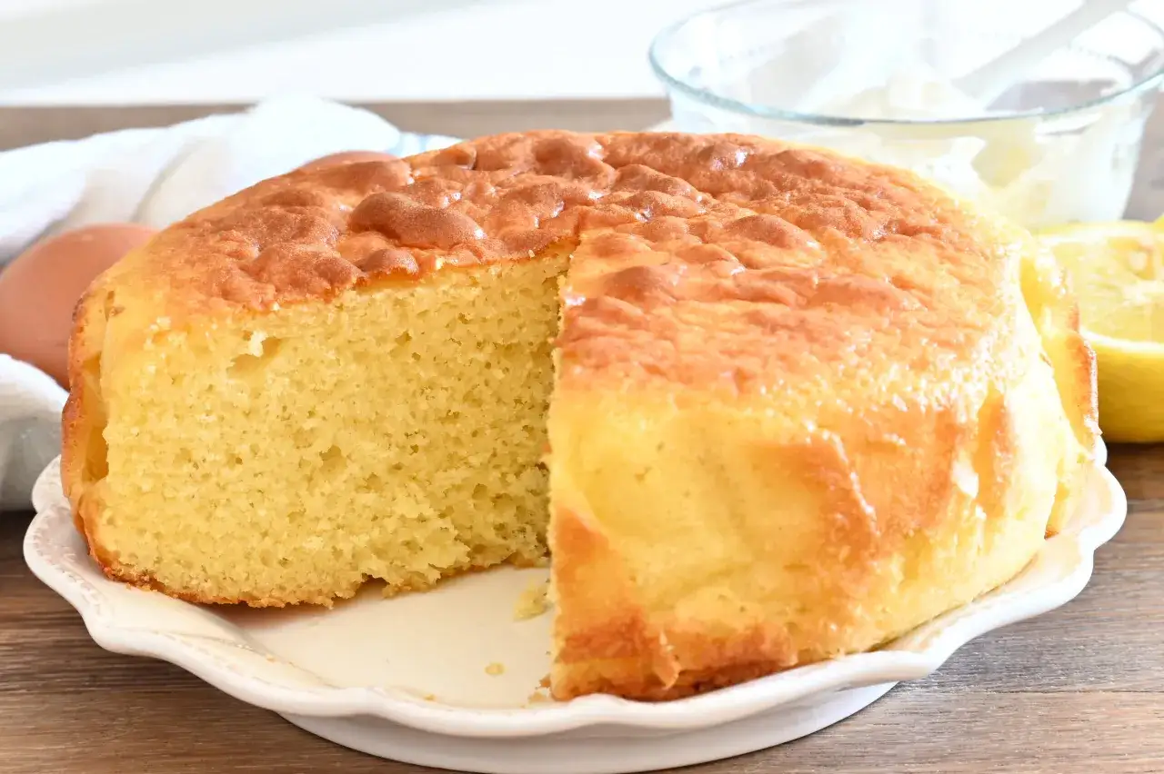 Una soffice torta per colazione, dorata e profumata, con una fetta mancante. Accanto, uova, limone e una ciotola di panna montata.