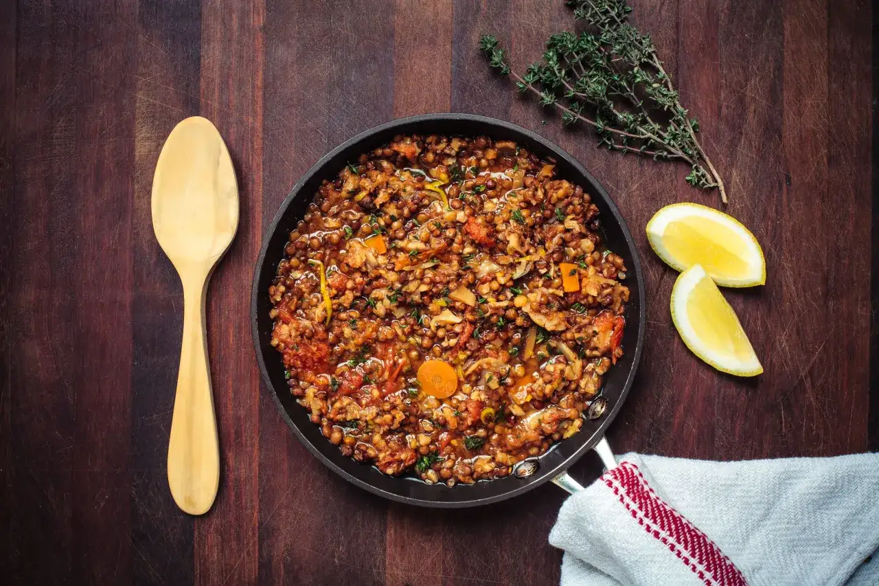 Un saporito ragù di lenticchie in padella, pronto per essere gustato. Accanto, limone fresco e un rametto di timo.