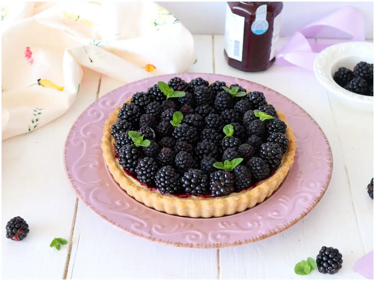 Deliziosa crostata di more, ricca di frutti succosi e guarnita con foglioline di menta, su un piatto rosa.