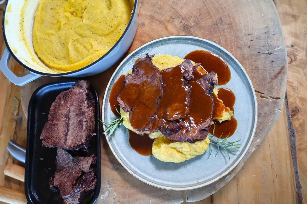 Un succulento brasato di manzo nappato con salsa, servito su un letto di polenta cremosa. Accanto, un pentolino di polenta e un piatto con altra carne.