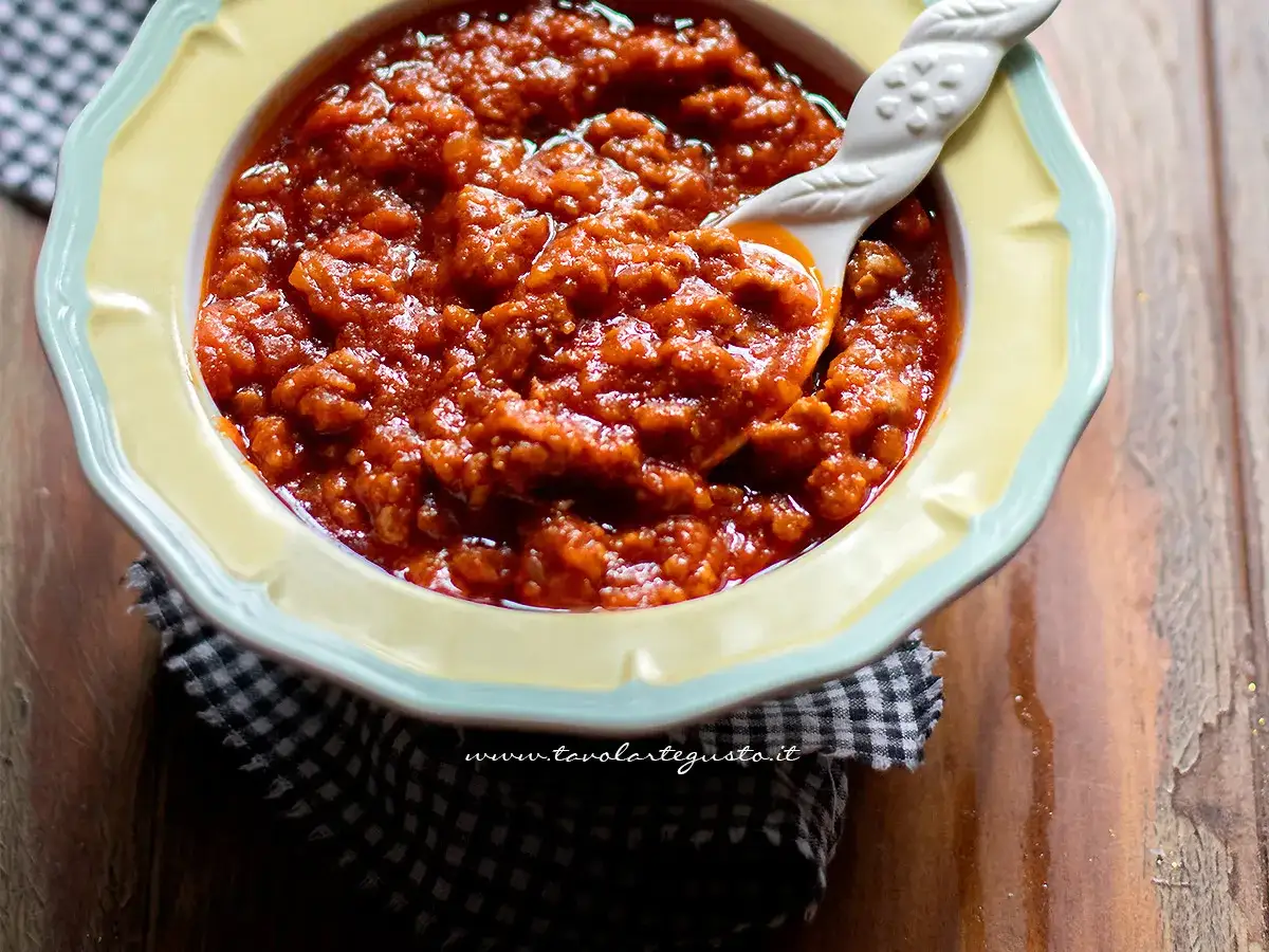 Un invitante sugo di salsiccia, denso e saporito, in una ciotola gialla con un cucchiaio decorato. Perfetto per un piatto di pasta.