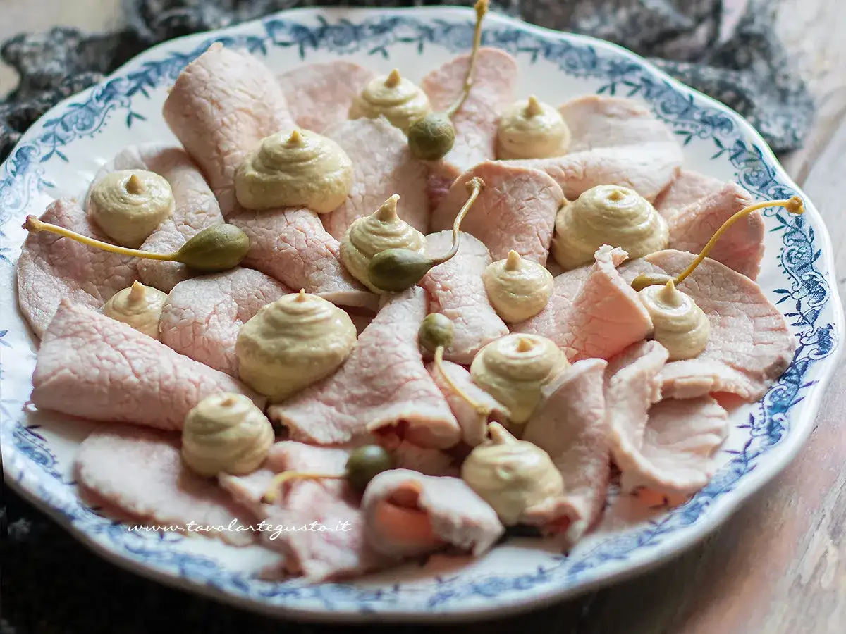 Vitello tonnato ricetta: fettine sottili di carne rosa, guarnite con salsa tonnata e capperi, su un piatto bianco con decori blu.