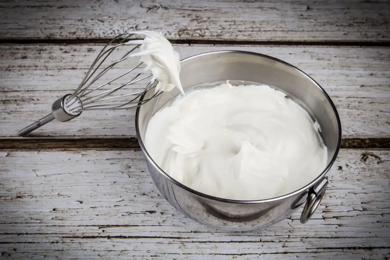 Frusta da cucina con panna da montare soffice e cremosa in una ciotola d'acciaio su un tavolo di legno rustico.