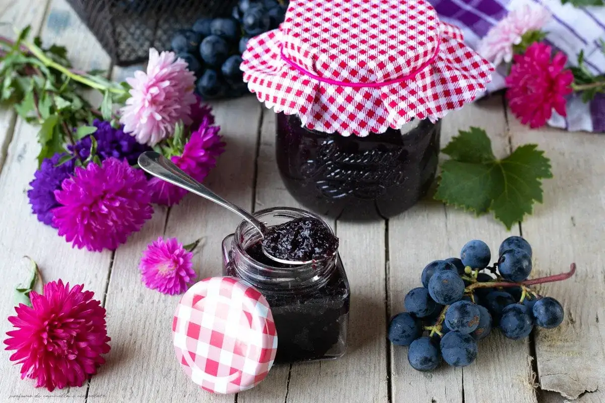 Un vasetto aperto con cucchiaino pieno di marmellata uva fragola, circondato da grappoli d'uva scura e fiori colorati su un tavolo di legno.