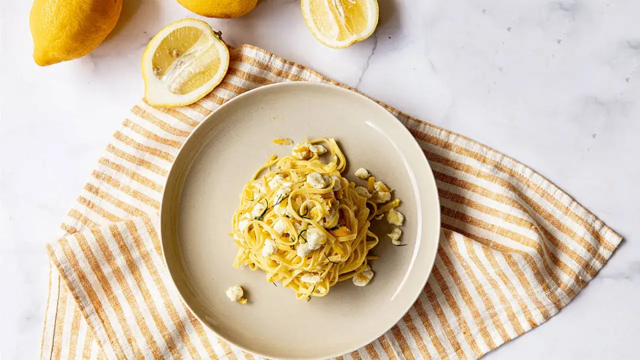 Un piatto di tagliolini al limone, fresco e profumato, con scorzette e cubetti di formaggio. Accanto, limoni freschi e un tovagliolo a righe.