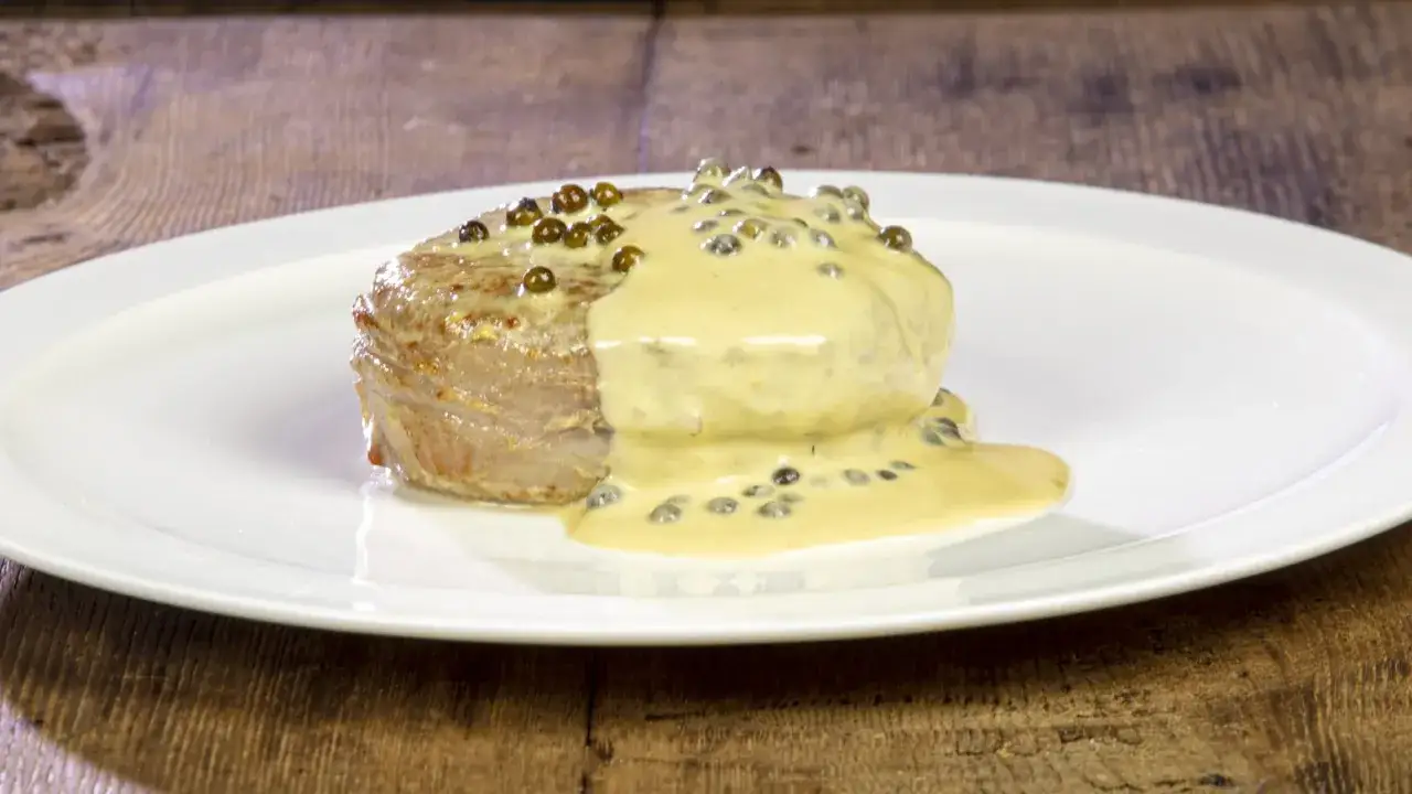 Un succulento filetto al pepe verde, nappato con una cremosa salsa, servito su un piatto bianco.