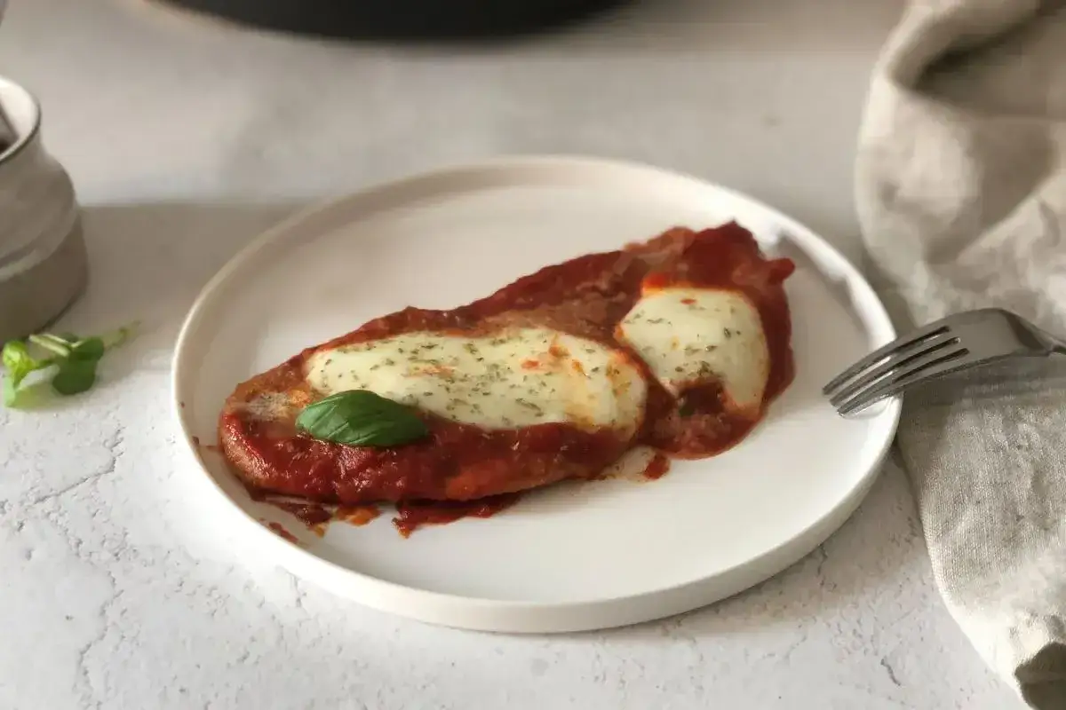 Scaloppine alla pizzaiola su un piatto bianco, con mozzarella filante, salsa di pomodoro e una foglia di basilico.