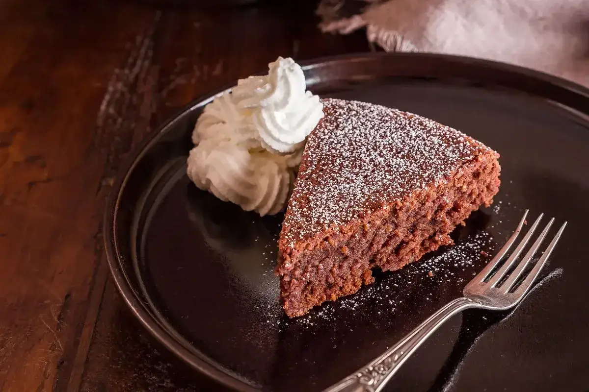 Una fetta di torta caprese bimby, soffice e al cioccolato, spolverata di zucchero a velo, servita con panna montata e una forchetta su un piatto scuro.