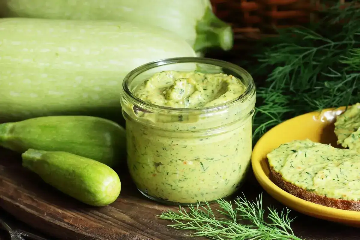Pesto di zucchine Bimby in un vasetto, con zucchine fresche e aneto. Perfetto spalmato su pane tostato.
