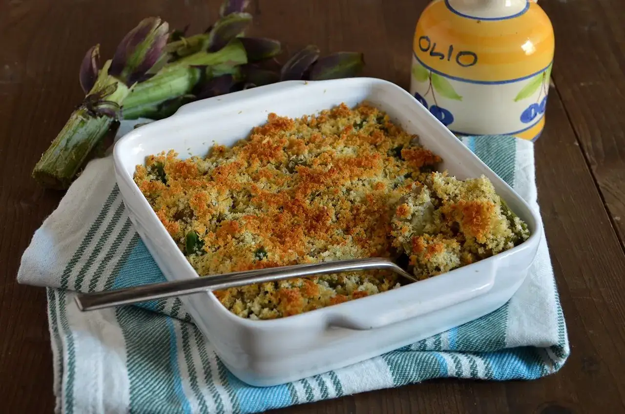 Piatto bianco con carciofi al forno morbidi, croccanti e dorati. Accanto, un mazzo di carciofi freschi e una bottiglia di olio.