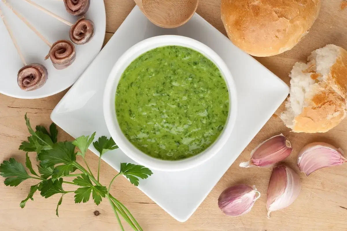 La ricetta salsa verde &egrave; pronta! Un ciotola di salsa verde brillante, aglio, prezzemolo e acciughe arrotolate.