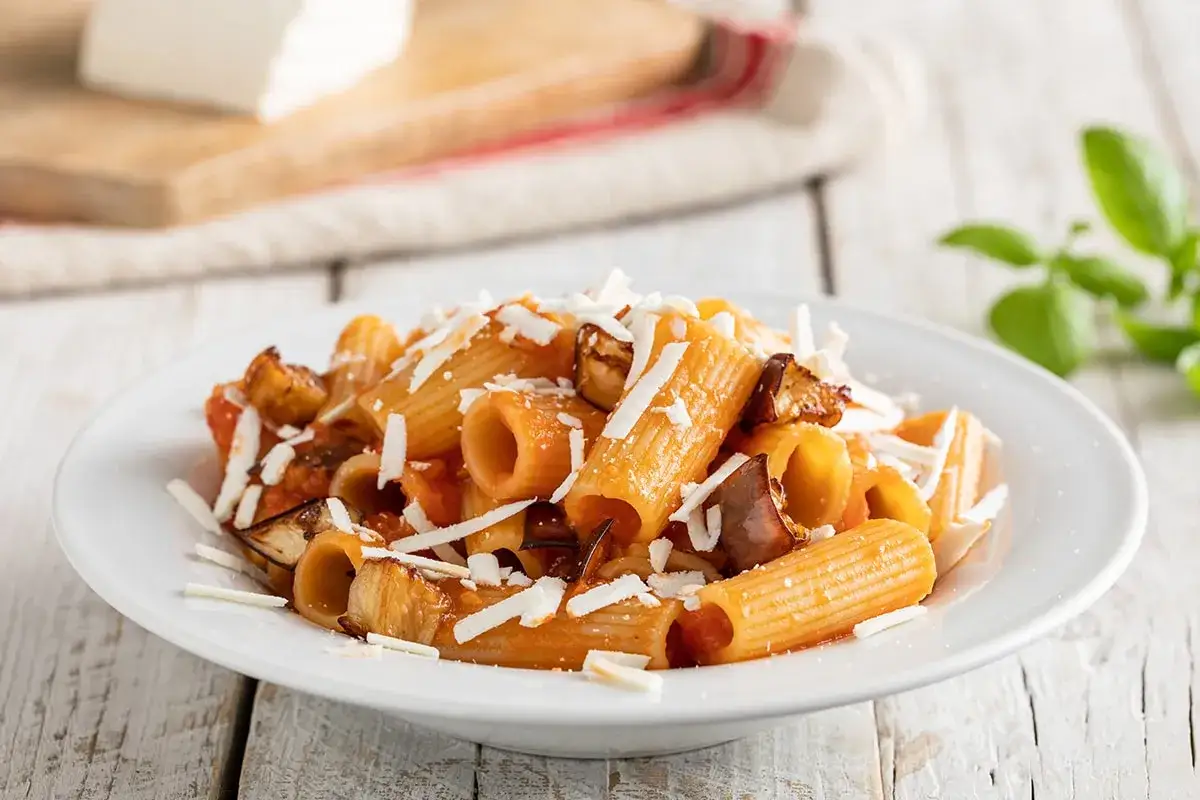 Piatto di pasta alla norma ricetta con melanzane fritte, pomodoro e ricotta salata grattugiata.